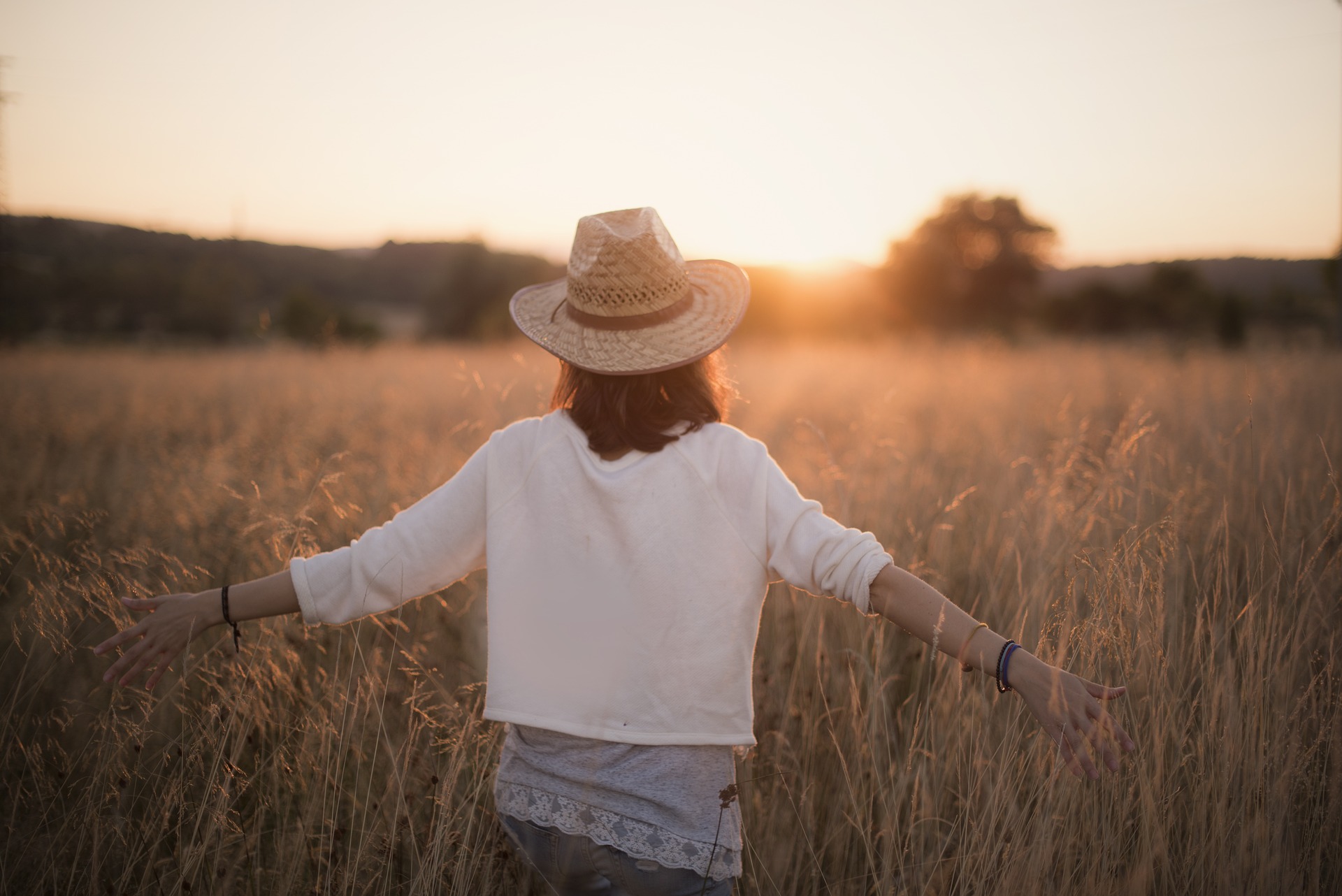 Garota vestindo blusa branca e chapéu de palha, andando de braços abertos num campo de capim alto. Sol poente ao fundo