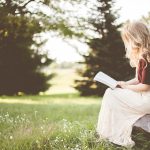 Garota de saia longa branca e camiseta preta sentada em um tronco num campo lendo um livro