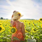 Mulher com vestido laranja e chapéu de palha andando em um campo de girassóis.