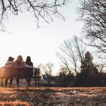 Três amigas sentadas em um banco numa paisagem típica de outono