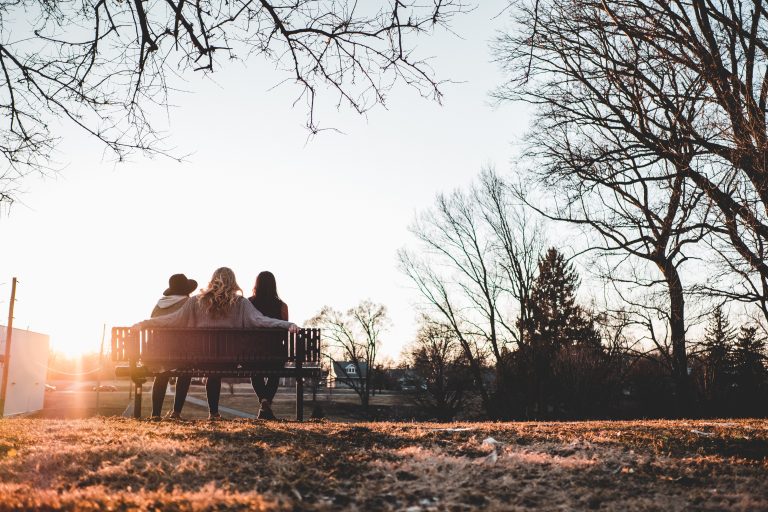 Três amigas sentadas em um banco numa paisagem típica de outono