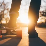 pernas de homem em jeans, com um pé em cima de um skate na rua. Sol brilhando ao fundo.