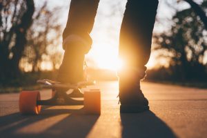 pernas de homem em jeans, com um pé em cima de um skate na rua. Sol brilhando ao fundo.