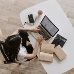 vista de cima uma mulher trabalhando no computador, com livros de um lado e celular e xícara de café do outro.