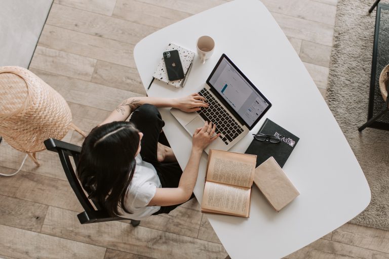 vista de cima uma mulher trabalhando no computador, com livros de um lado e celular e xícara de café do outro.