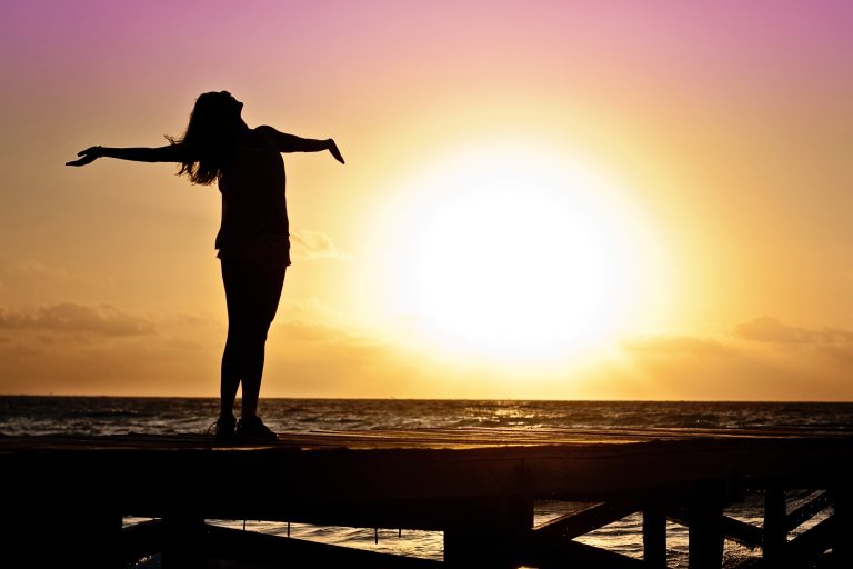 mulher de braços abertos em píer na praia, pôr do sol ao fundo