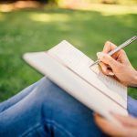 Mulher sentada na grama, apoiando um caderno nos joelhos dobrados, escrevendo