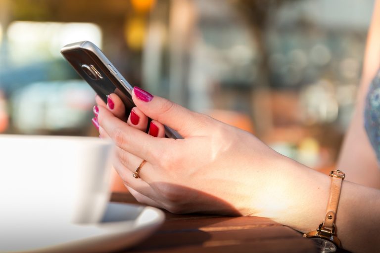 mulher com unha pintada de vermelho segurando um celular sentada à mesa com xícara de café