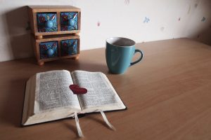 biblia aberta sobre uma mesa, xícara ao lado, gavetinhas azuis ao fundo